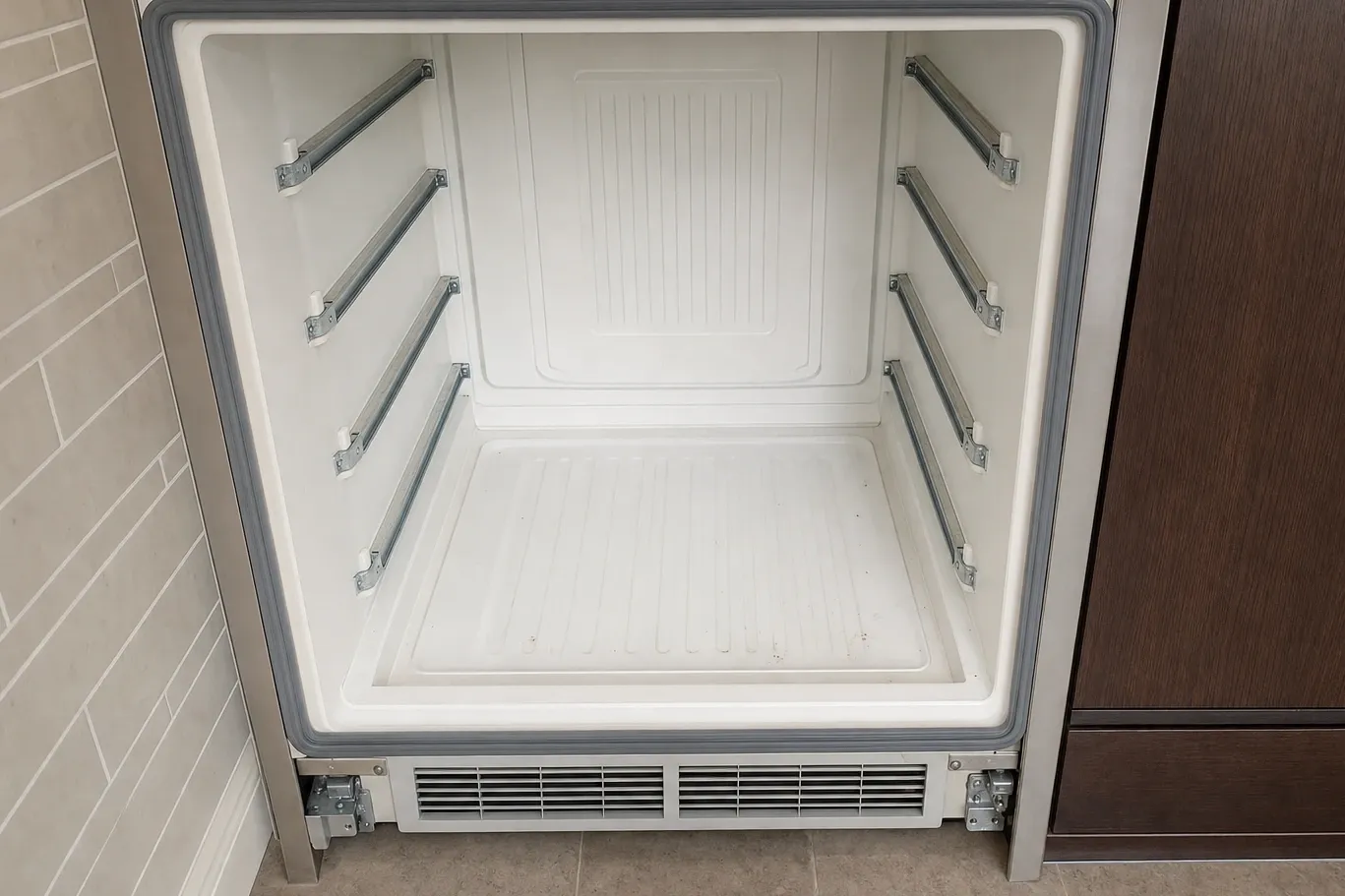 Undercounter refrigerator built into cabinetry, door open on an empty white interior with shelf rails and a base vent grille